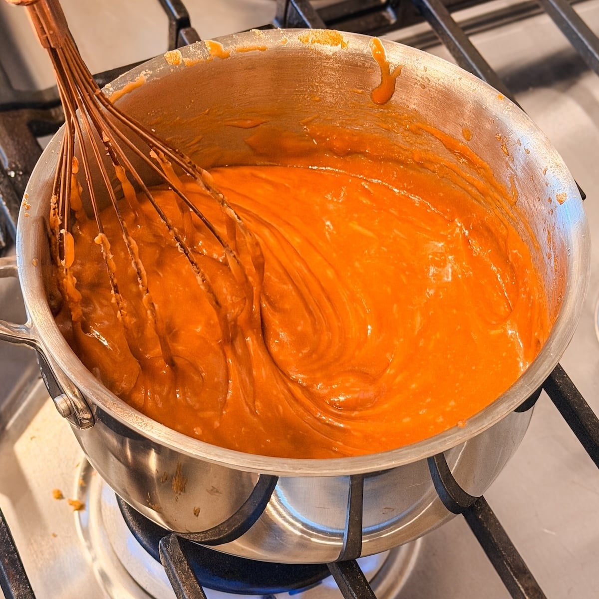 A whisk stirs thick, orange sauce in a metal pot on a gas stove burner, with sauce splatters visible on the pots rim. It is a deep golden brown color