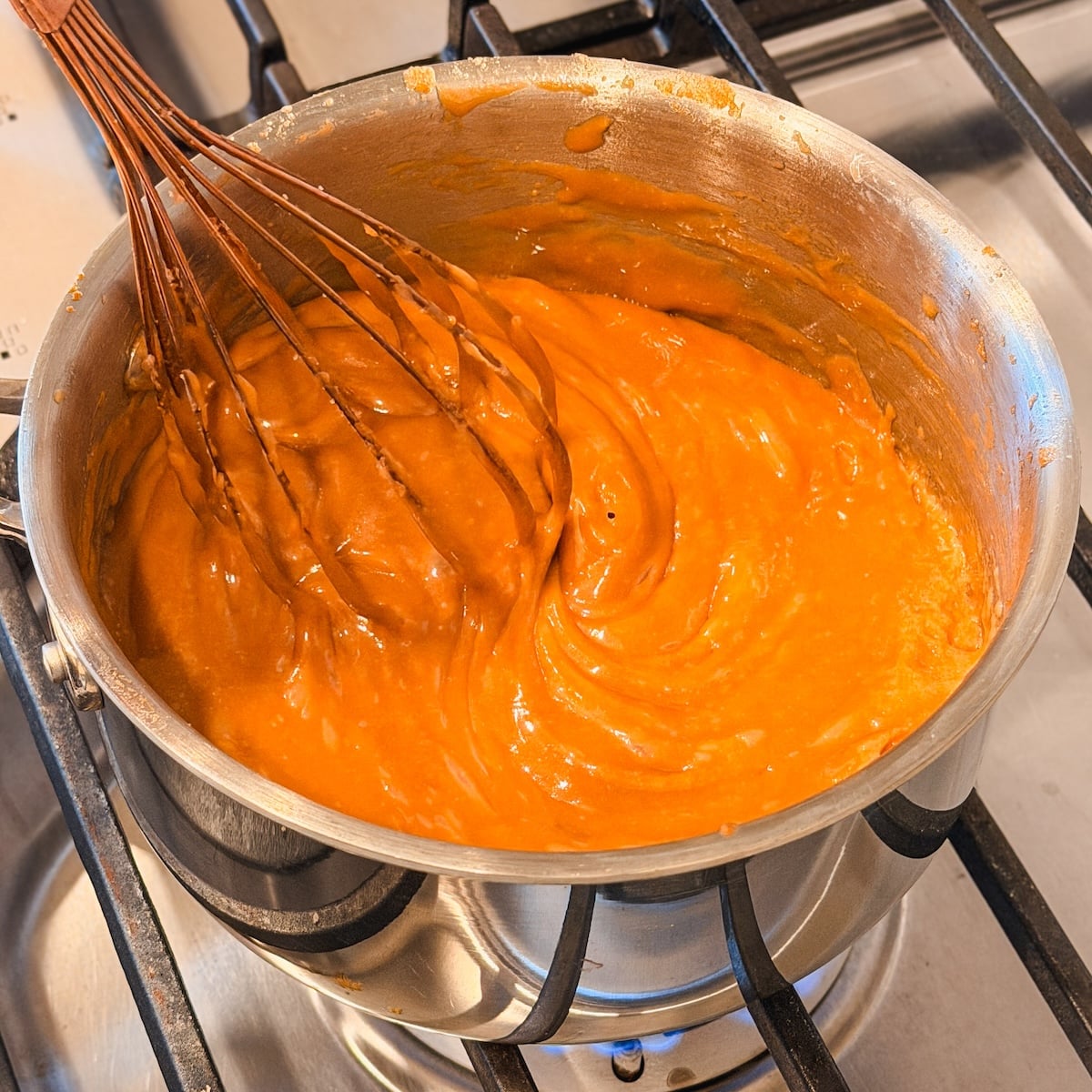 A metal saucepan containing thick orange sauce is being whisked on a gas stove. The sauce is smooth and glossy, and the whisk is partially coated with it. The stove burners are visible below the pot.