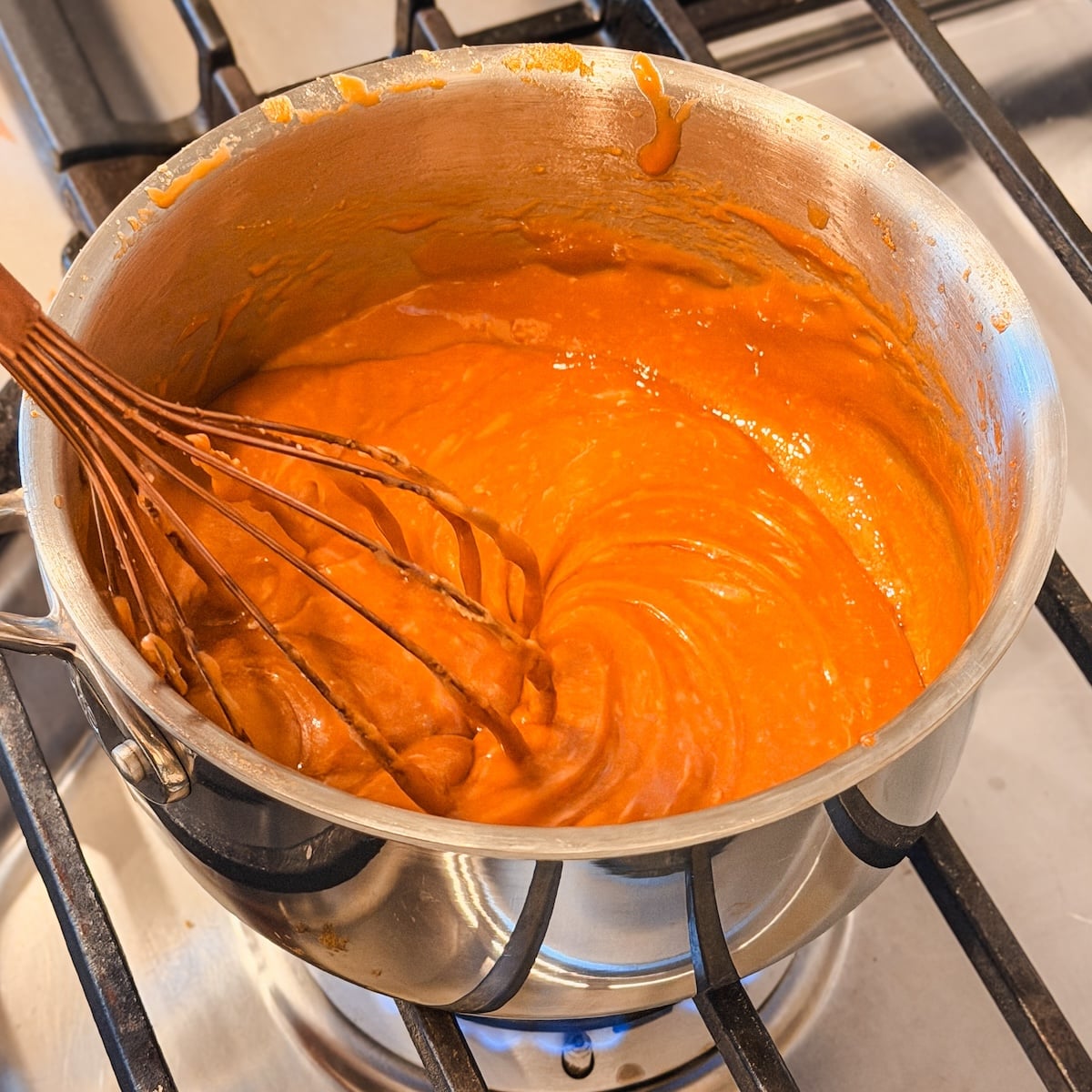 A metal pot on a stove contains thick orange sauce being whisked. The sauce has a creamy texture, and the whisk is partially covered in the mixture, showing it being stirred. The sauce seems to be gliding on the surface of the pan