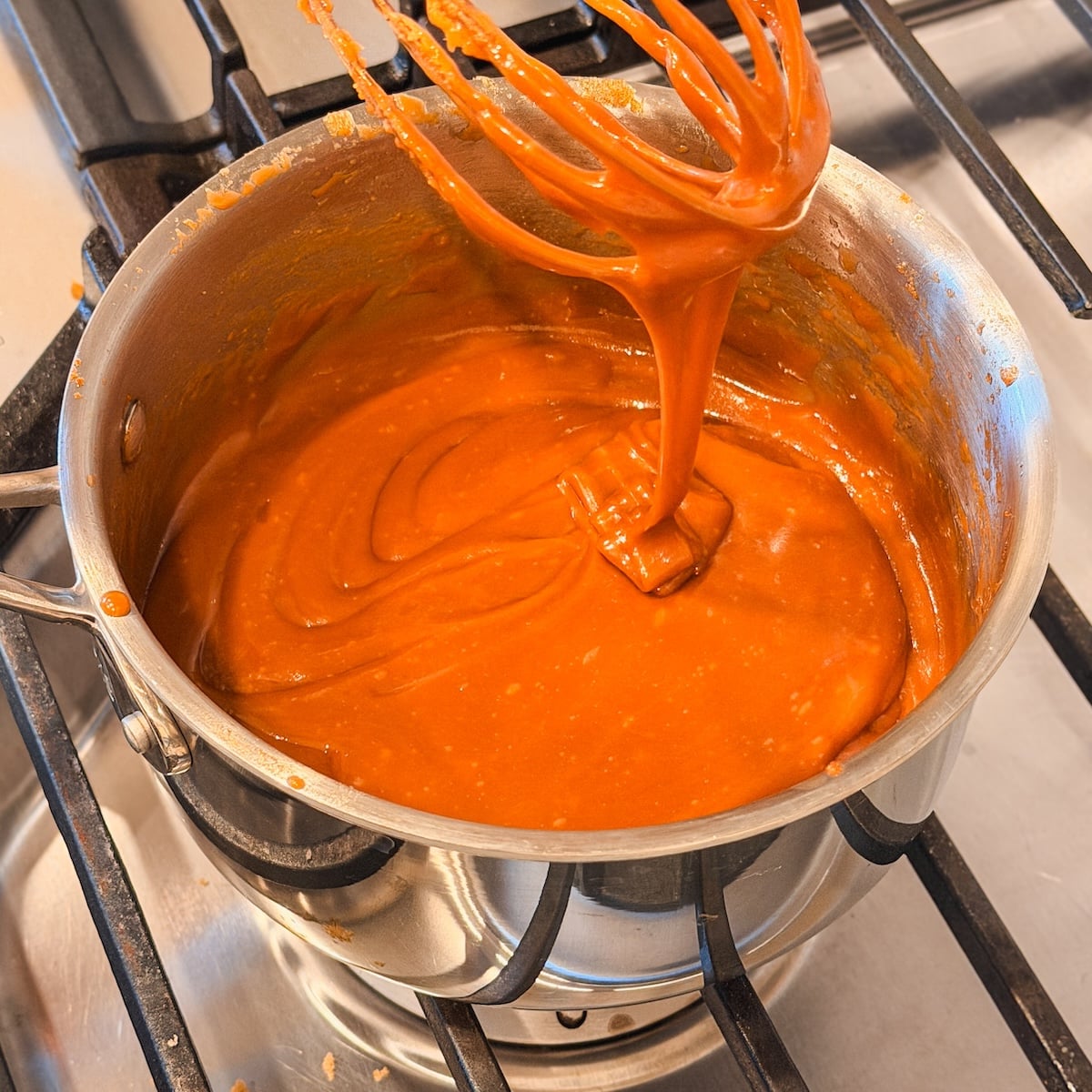 A whisk lifts thick, creamy toffee sauce from a stainless steel pot on a stove, with toffee drips visible.
