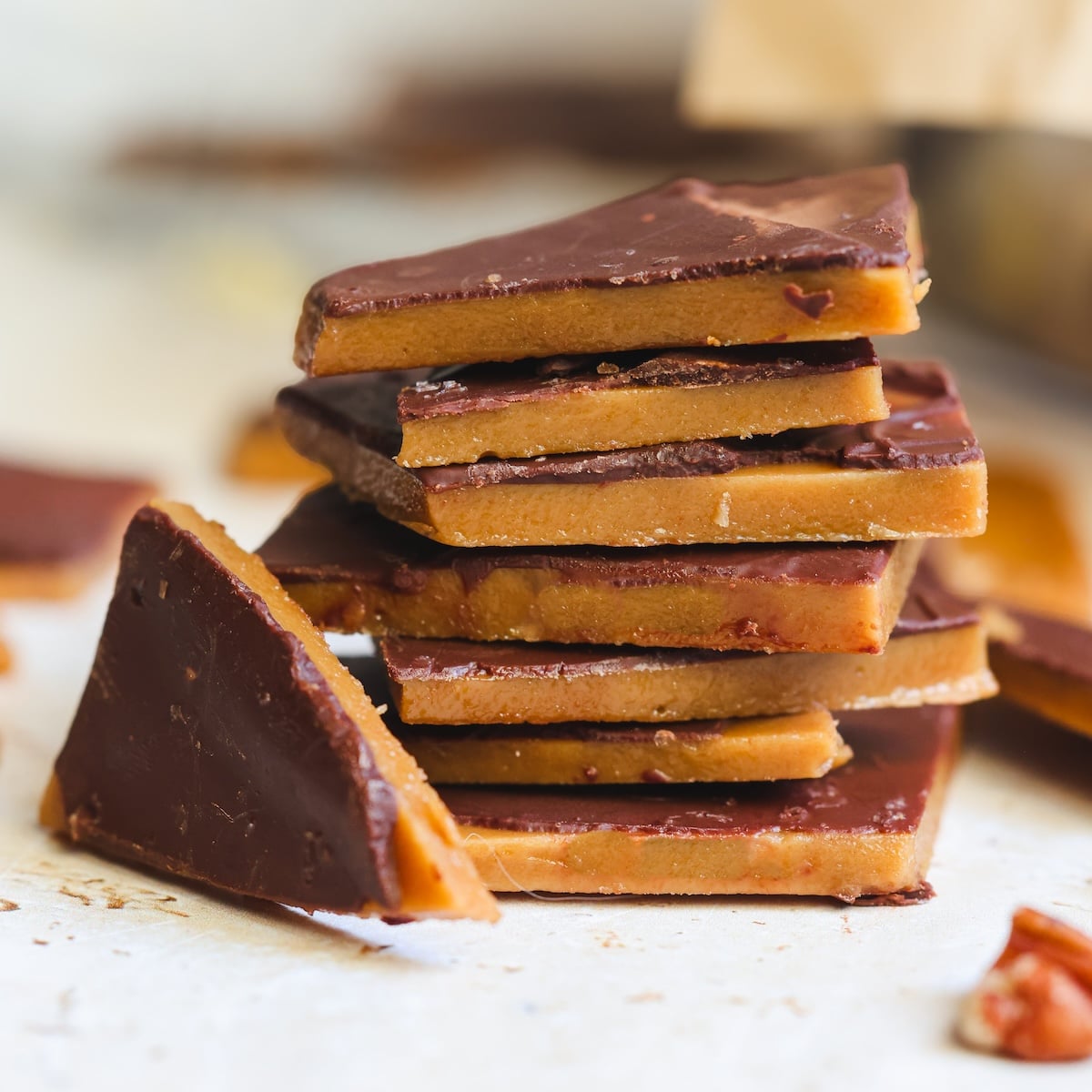 A stack of homemade toffee pieces coated with chocolate, with a few extra pieces scattered around on a light surface.