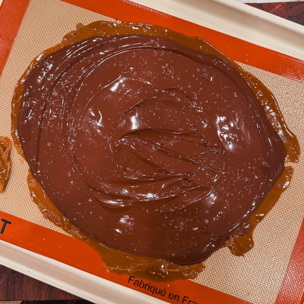 Melted chocolate spread over a layer of toffee on a silicone baking mat, with a sprinkle of sea salt on top.