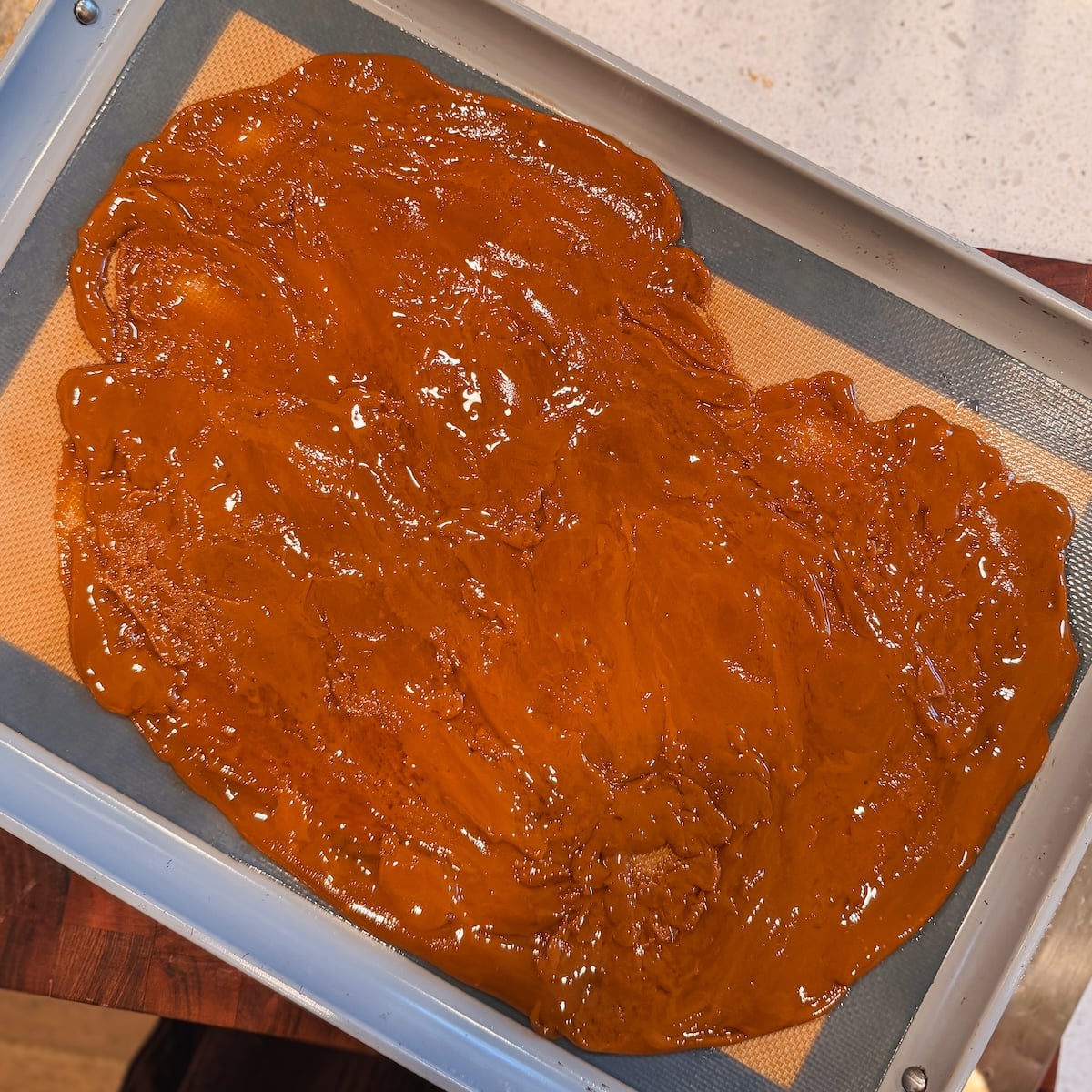 A baking tray with a silicone mat holds a large sheet of shiny, golden-brown toffee spread thinly across the surface.