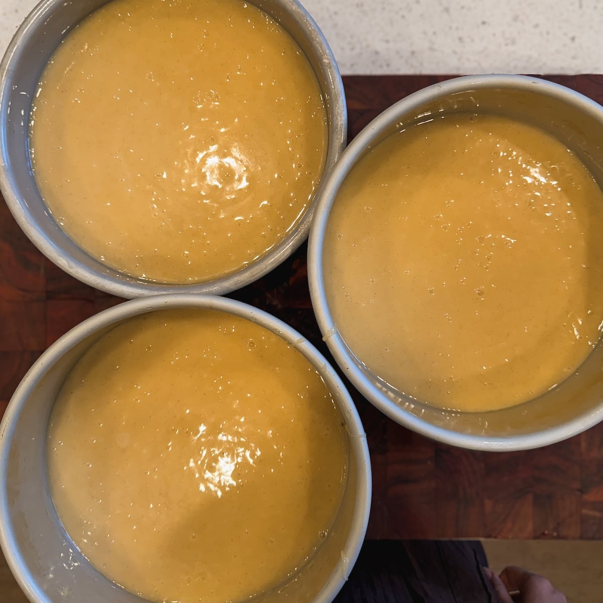 Three round cake pans filled with vanilla cake batter sit on a wooden surface, viewed from above.