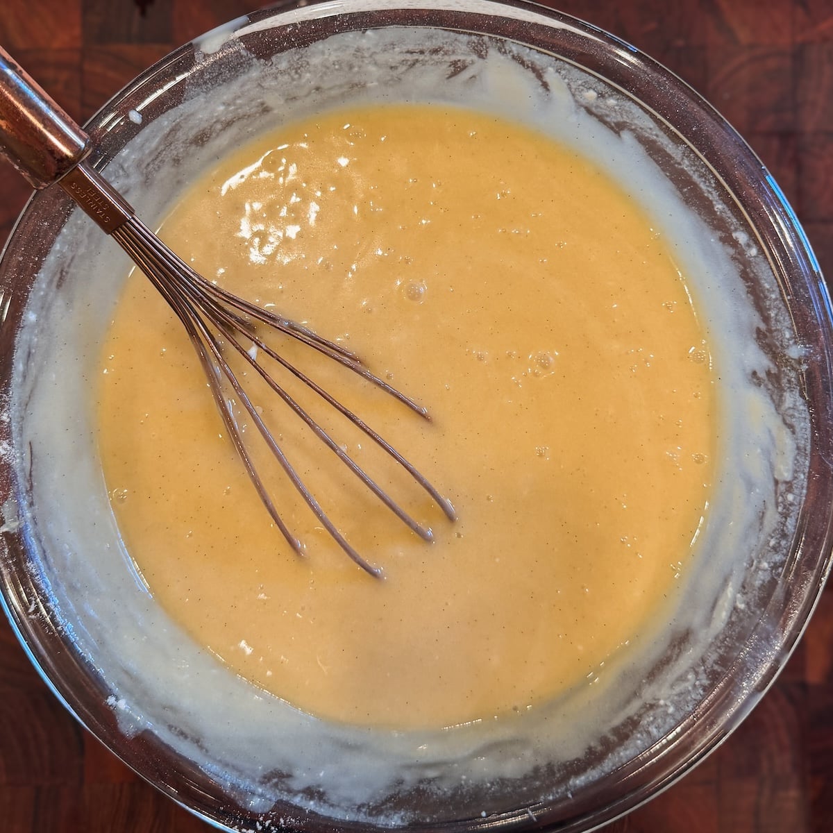 A metal whisk rests in a glass bowl filled with smooth, pale yellow batter. The bowls rim has batter smeared on it, and the surface below is a dark wooden countertop.