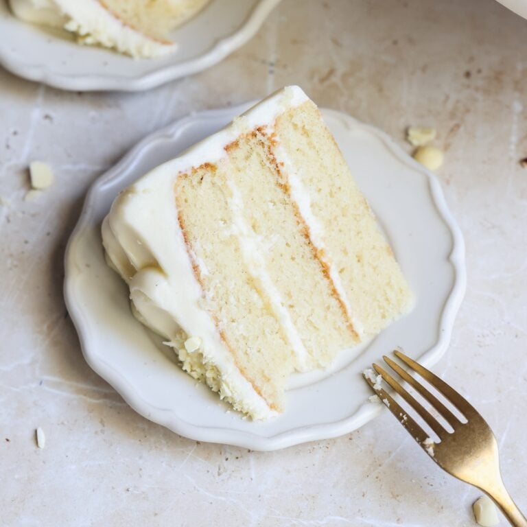 A slice of three-layer vanilla cake with white frosting sits on a white plate. A gold fork rests beside the cake. The background is a light marble surface.