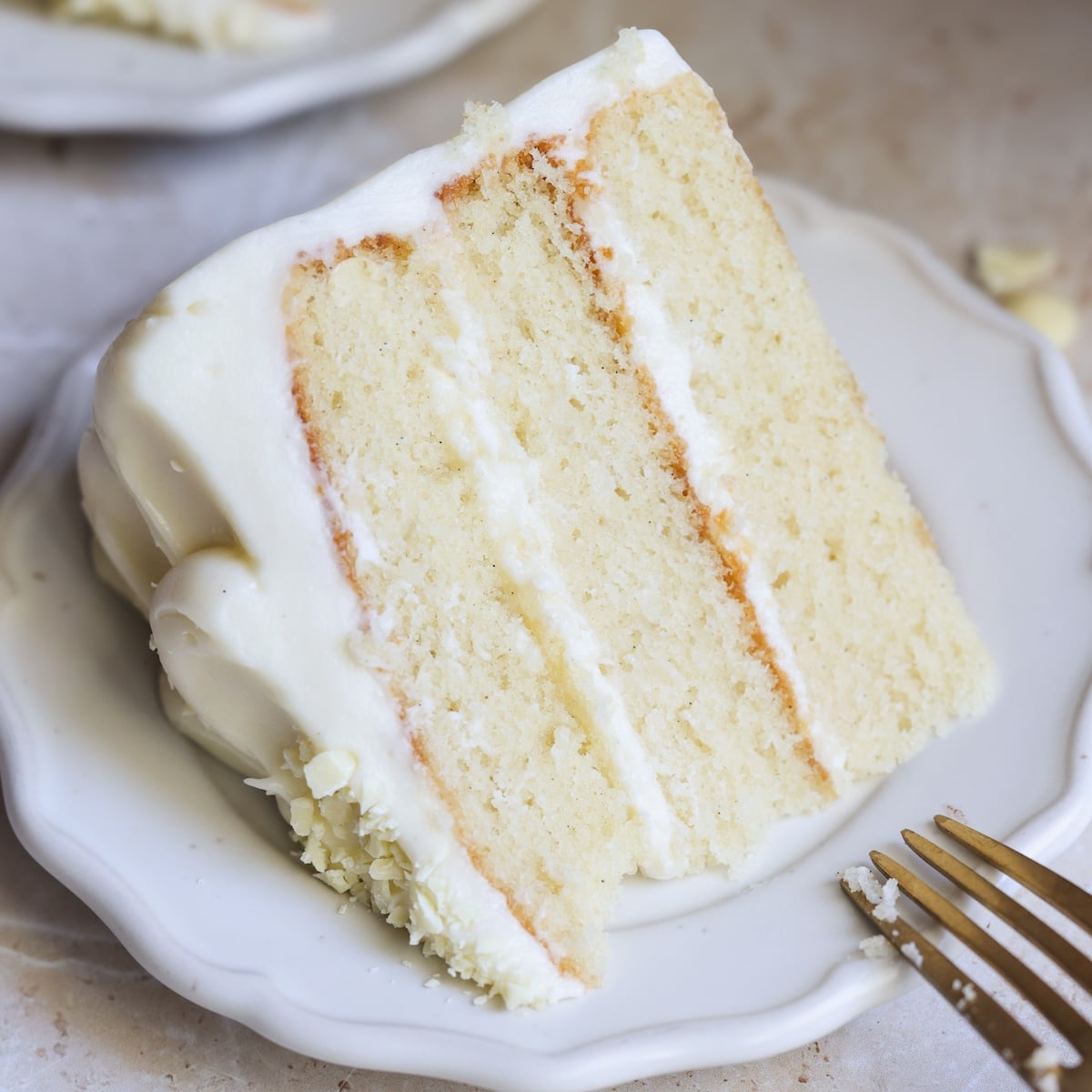 A slice of three-layer vanilla cake with white frosting sits on a white plate. A fork rests nearby. The cake looks moist and fluffy, with smooth, creamy icing between the layers and on the outside.