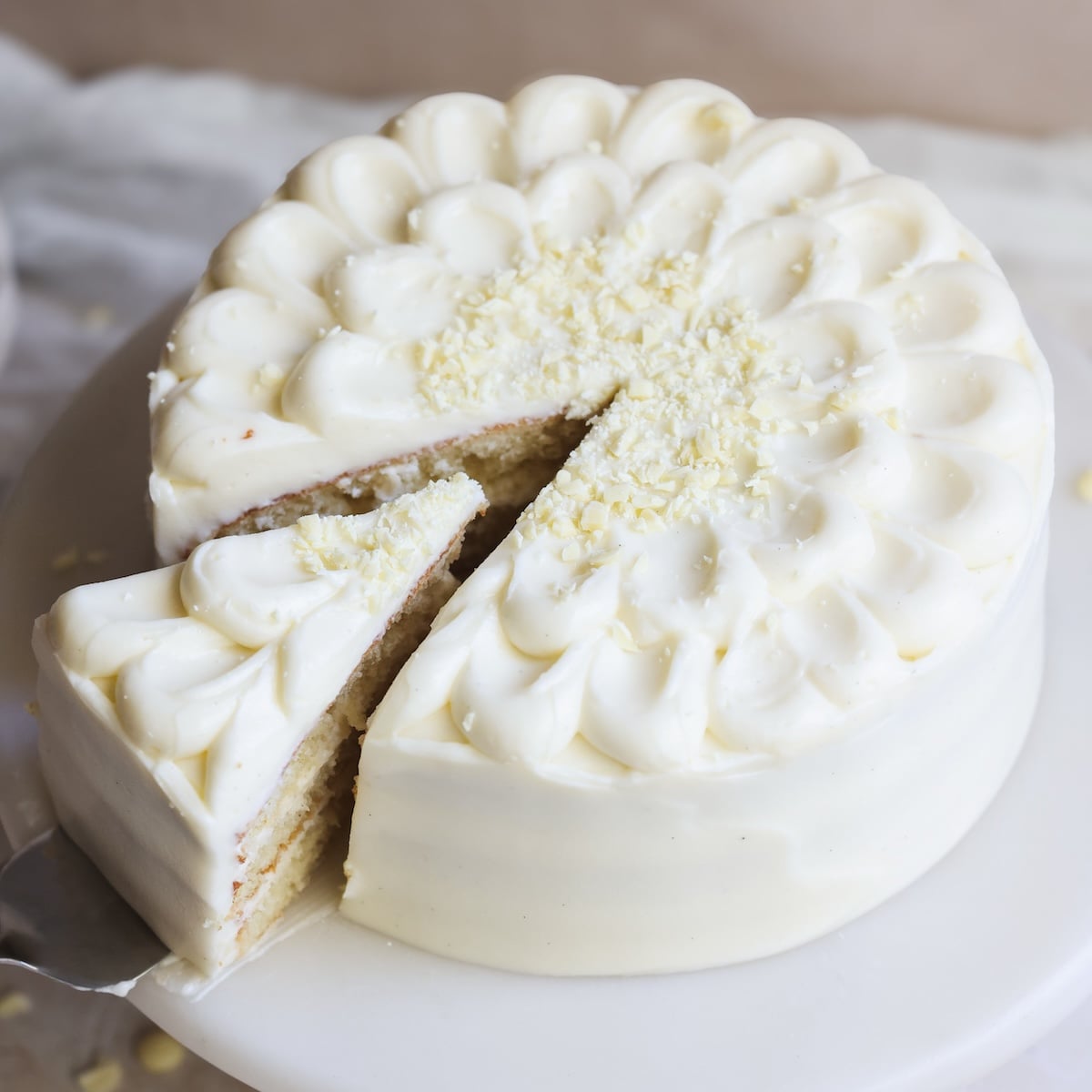 A round white frosted vanilla cake with a slice being lifted out. The cake is topped with piped frosting and white chocolate shavings.