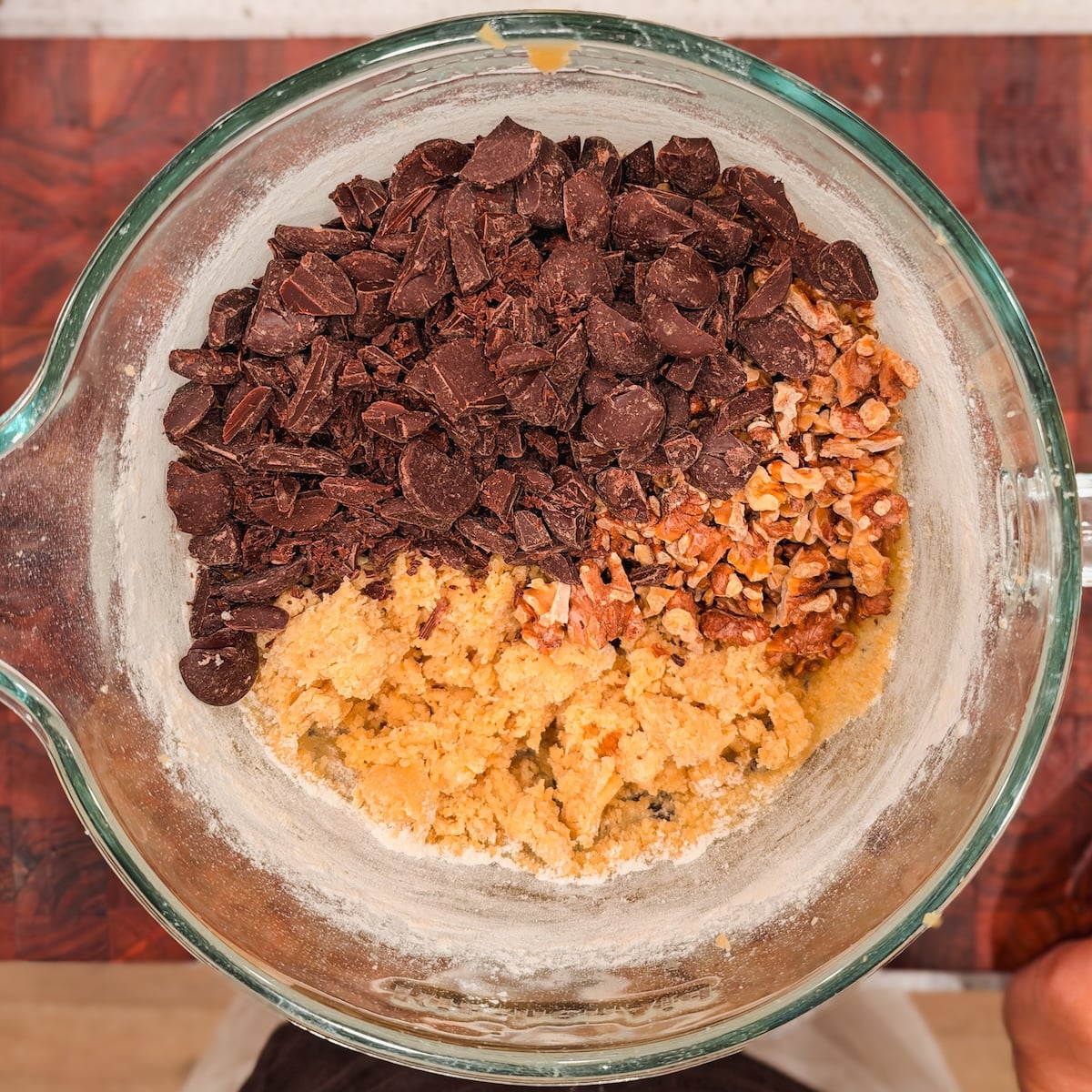 A clear glass bowl contains cookie dough topped with a pile of chopped chocolate chunks and chopped walnuts