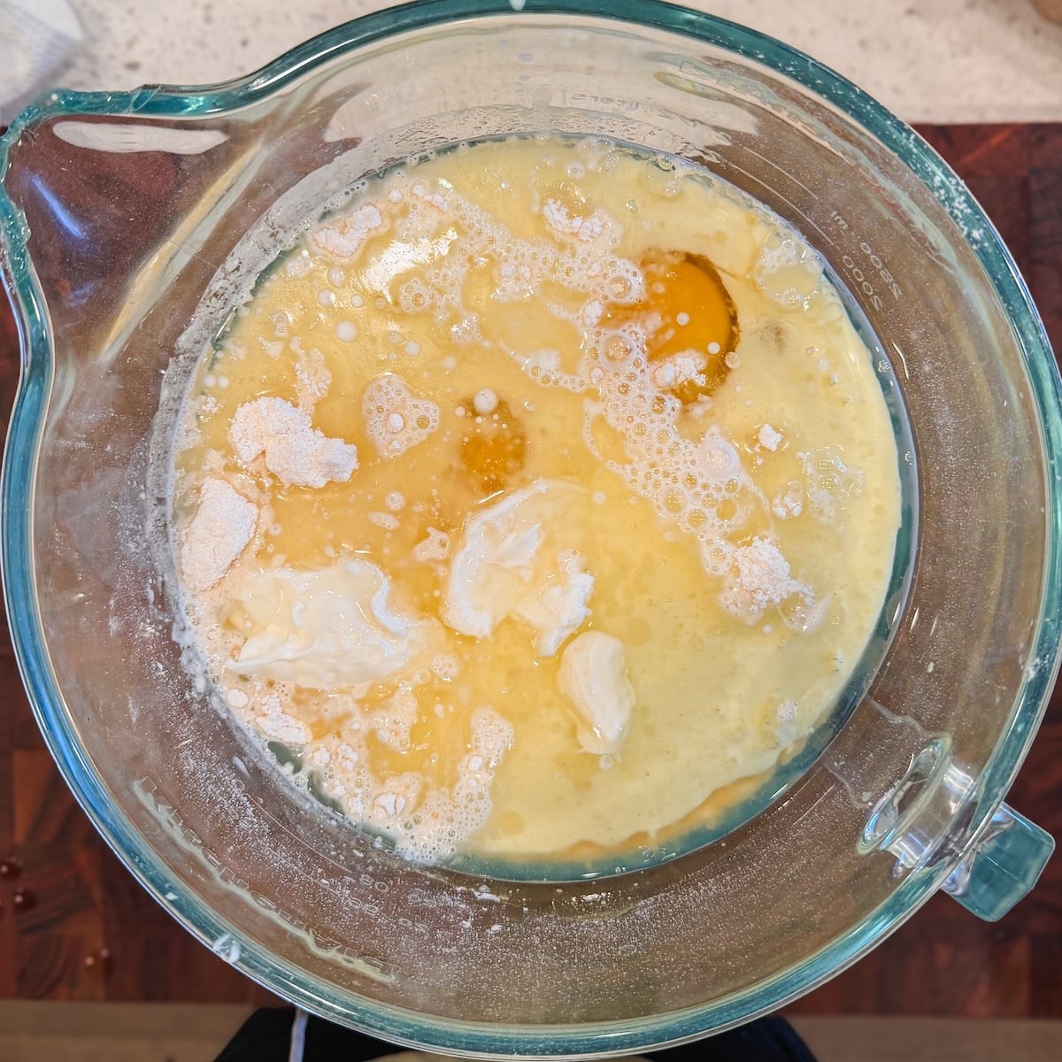 A glass bowl contains cracked eggs, flour, and other baking ingredients being mixed, seen from above on a kitchen countertop.