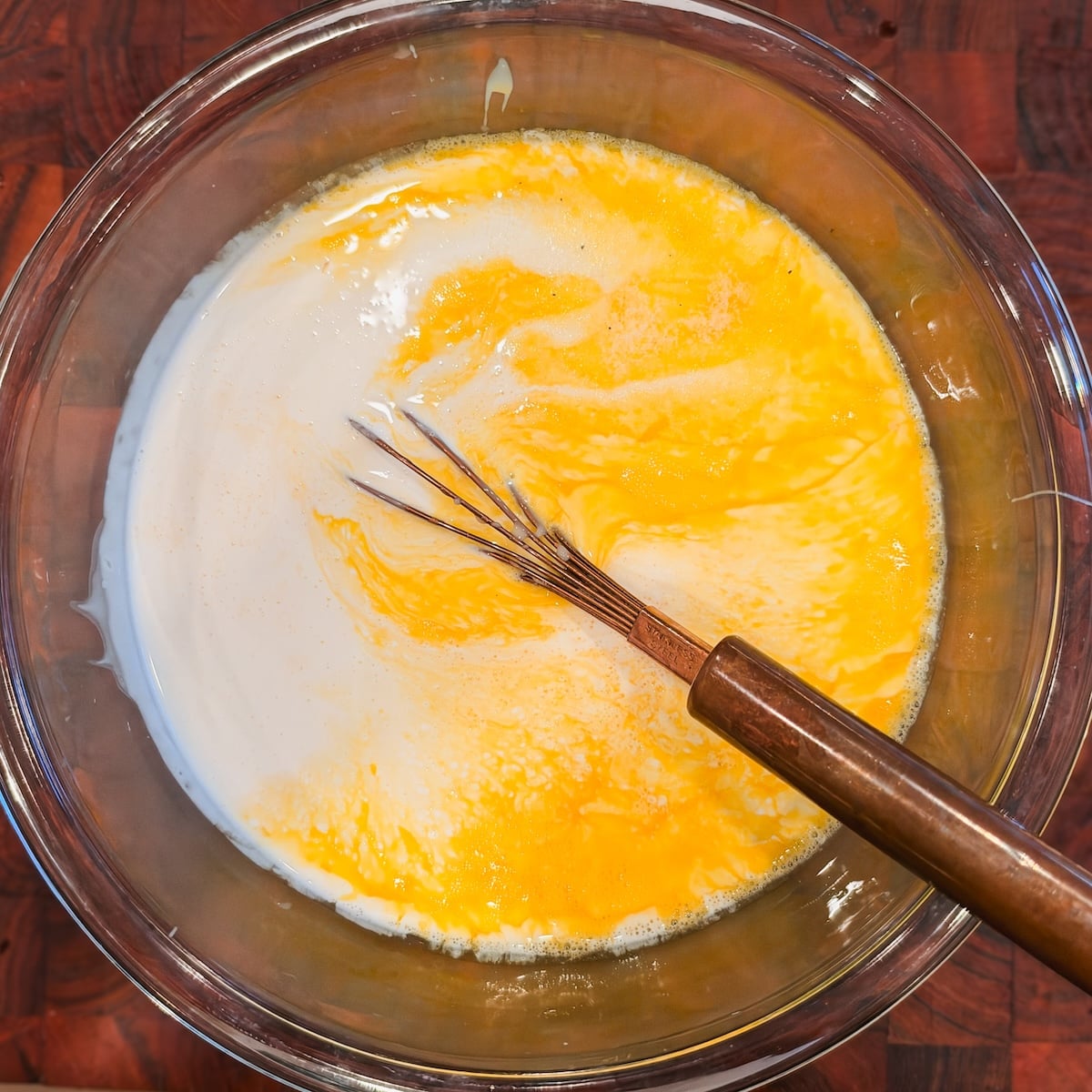 A glass bowl containing eggs, cream and sweetened condensed milk being mixed with a metal whisk, on a wooden surface.