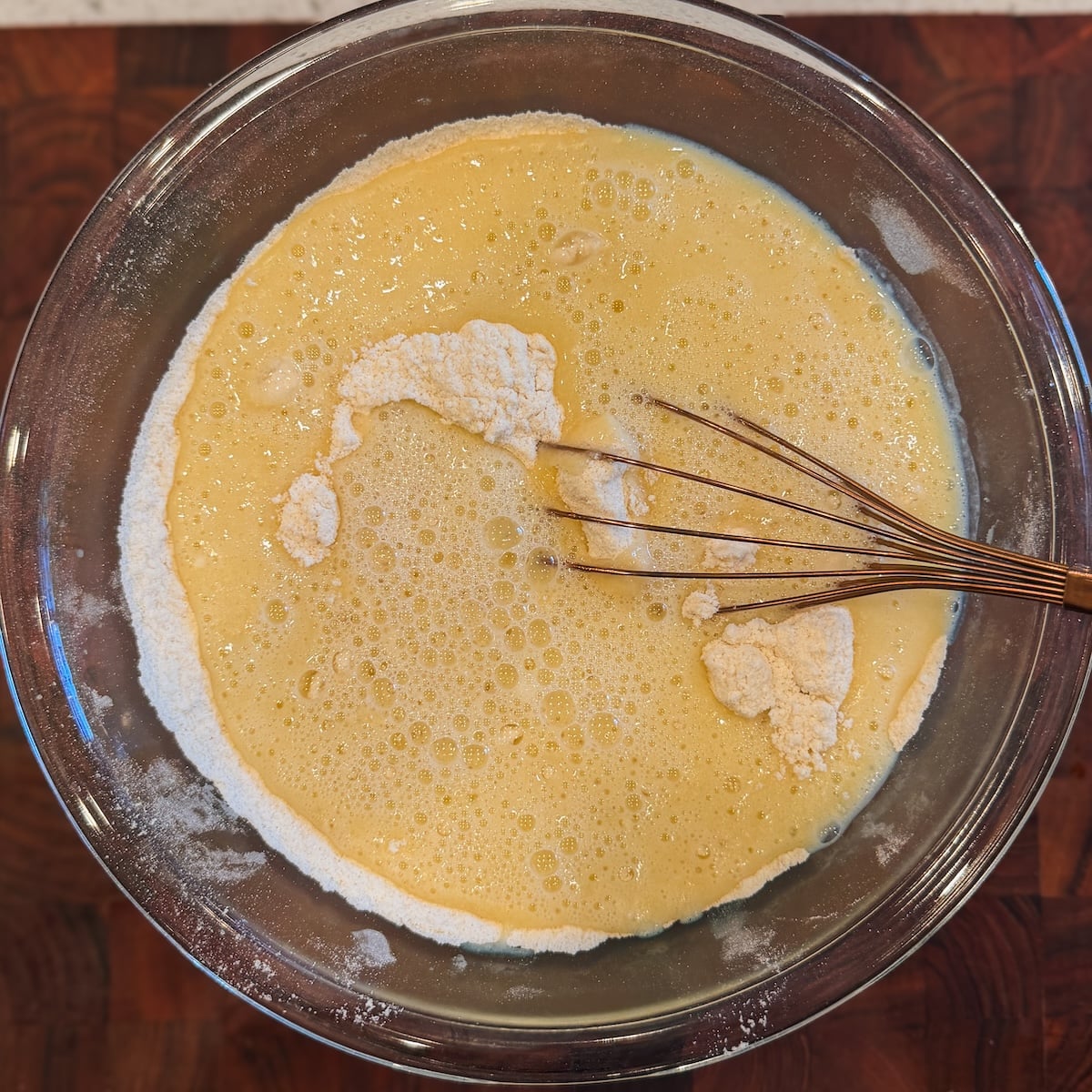 A glass bowl contains flour and a yellow batter mixture being mixed with a whisk, placed on a dark wooden surface.